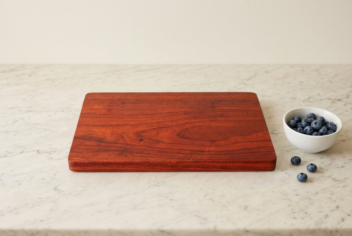 Padauk wood cutting board on marble counter beside small white bowl of fresh blueberries, simple natural kitchen light