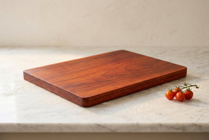 Solid African padauk cutting board low angle on white marble showing crisp edge and board thickness with small vine of cherry tomatoes
