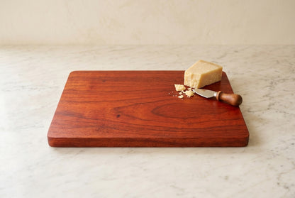 Padauk cutting board with small wedge of aged parmesan, crumbs and cheese knife on one corner, white marble counter
