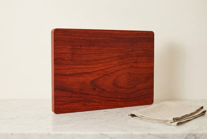 Single slab padauk cutting board standing upright showing full red orange grain face with folded linen tea towel on marble