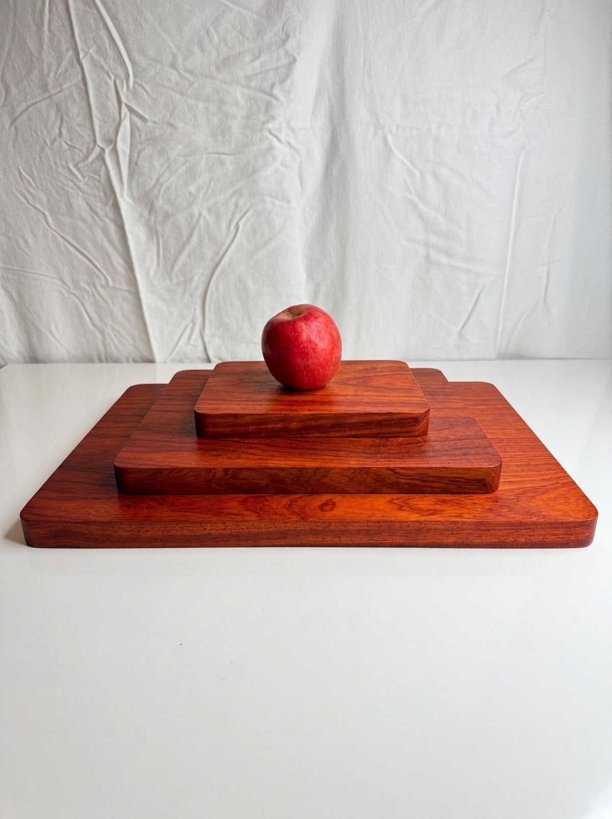 Three padauk cutting boards small medium and large stacked with a red apple for size comparison on white surface
