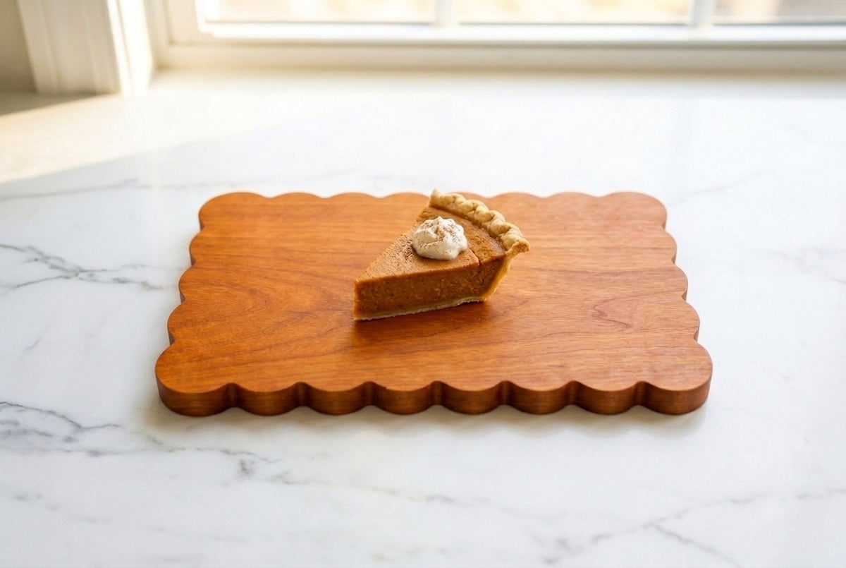 Padauk scalloped charcuterie board with a single slice of pumpkin pie and whipped cream on white marble counter, warm window light