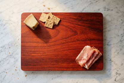 Overhead padauk cutting board minimal charcuterie prosciutto aged cheese crackers sparse elegant on white marble counter