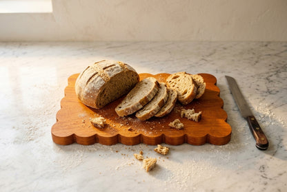 Padauk scalloped board with sliced artisan sourdough loaf and bread knife warm kitchen window light with flour dust crumbs