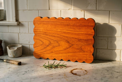 Padauk scalloped charcuterie board standing upright against subway tile showing wavy hand cut edges and vivid orange grain